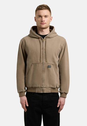 Mand iført lys brun hættetrøje med lynlås og forlommer samt sorte bukser, stående foran en ensfarvet hvid baggrund.