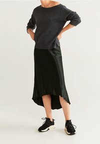 Personne portant un pull gris foncé, une jupe satinée noire asymétrique et des baskets noires et blanches, debout les mains sur les hanches.