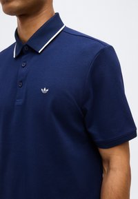 Άνδρας με μπλε σκούρο πόλο πουκάμισο με λευκή περιγραφή στο γιακά και κεντημένο λογότυπο στο αριστερό στήθος.