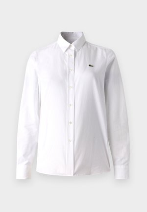 Hvit skjorte med knapper laget av bomull, med klassisk krage, lange ermer, og en liten grønn logo på venstre bryst.