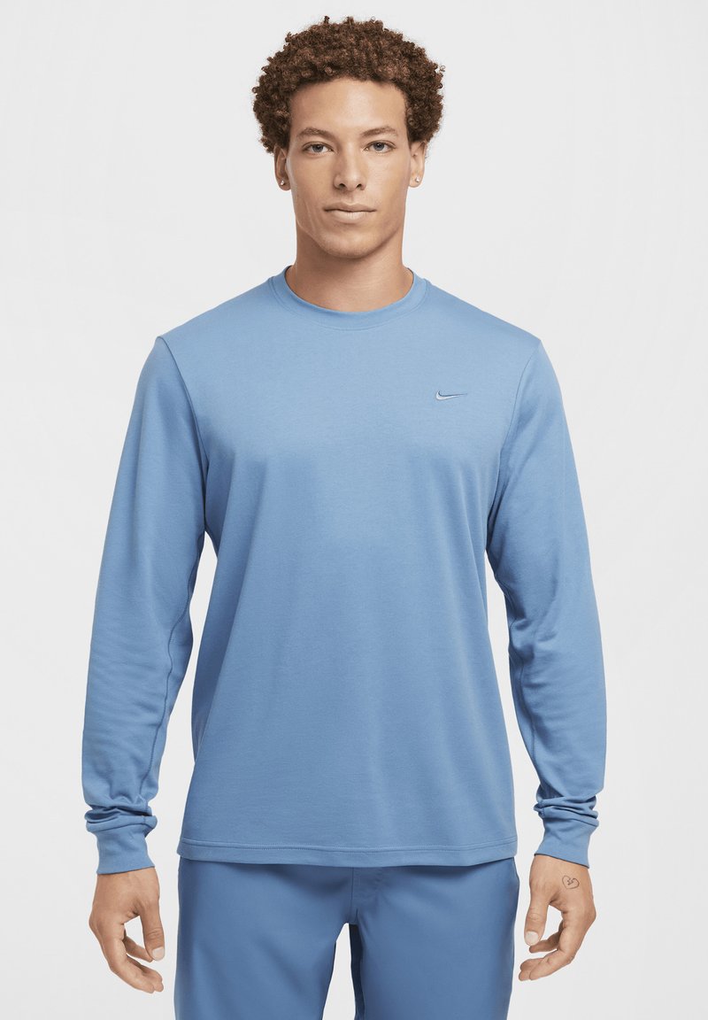 Mężczyzna z kręconymi włosami, ubrany w jasnoniebieską koszulkę z długim rękawem marki Nike oraz pasujące spodnie, stoi na tle białej, gładkiej ściany.