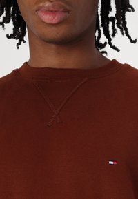 Felpa di cotone marrone con scollatura a girocollo e dettagli a punto croce. Include un piccolo logo ricamato in rosso, bianco e blu.