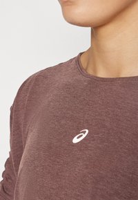 Camisa de manga larga de color burdeos hecha de una tela suave y texturizada. Presenta un pequeño logo blanco en el pecho y un sencillo cuello redondo.