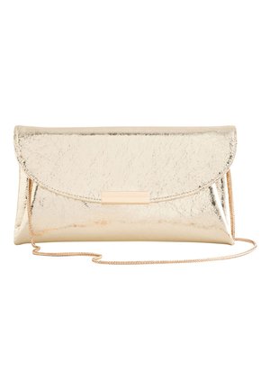 Frau mit schulterlangem braunem Haar trägt ein transparentes, mit Pailletten besetztes Oberteil und hat eine glänzende goldene Clutch über der Schulter.