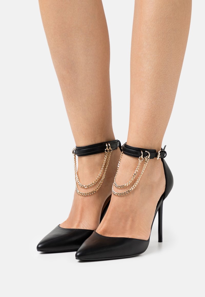 Chaussures noires à talons hauts à bout pointu avec brides de cheville ornées de détails en chaîne dorée, portées sur des jambes nues sur fond blanc.