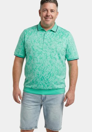 Mies hymyilee, hänellä on päällään turkoosinvärinen, lehtikuvioinen poolopaita ja vaaleansiniset farkkushortsit. Hän seisoo yksinkertaisen valkoisen taustan edessä.