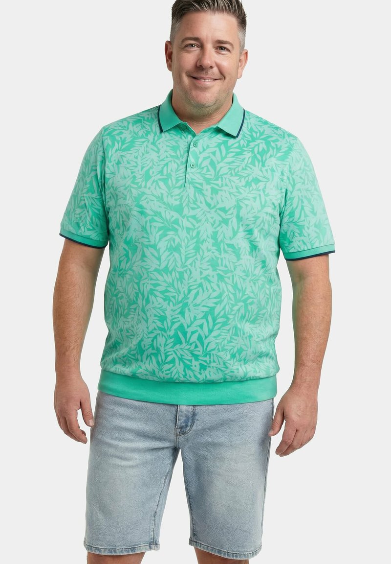 Mann lächelnd, trägt ein türkisfarbenes Poloshirt mit Blattmuster und hellblaue Jeansshorts, steht vor einem schlichten weißen Hintergrund.