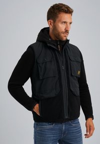 Chaleco negro con dos bolsillos frontales, hecho de un material texturizado, superpuesto a una sudadera con capucha de forro polar negra. Cierre de cremallera con detalle de logotipo.