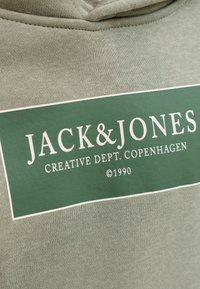 Alyvuogių spalvos audinys su lygiu paviršiumi, kuriame matomas iškilus baltas stačiakampis logotipas, ant kurio užrašyta "JACK & JONES" ir "CREATIVE DEPT. COPENHAGEN."