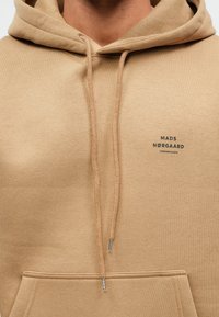 Κίτρινη κουκούλα με κορδόνια, απαλό υλικό, τσέπη καγκουρό και μικρό μαύρο λογότυπο "MADS NØRGAARD COPENHAGEN" μπροστά.