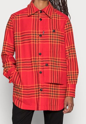Rood-oranje geruit shirt met lange mouwen, zwarte knopen, borstzak en kraag gedragen door een persoon met donker gevlochten haar.