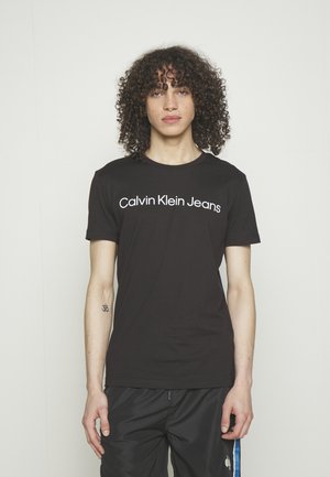Personne aux cheveux bouclés portant un t-shirt noir Calvin Klein Jeans et un pantalon de jogging noir avec un petit tatouage sur l'avant-bras gauche.
