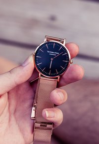 Okrągły czarny zegarek z różowo-złotą bransoletą mesh, z minimalistycznymi znacznikami godzin, nazwa marki "Victoria-Hyde London" umieszczona w pobliżu środka.
