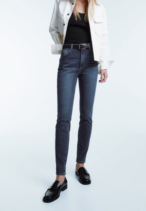 Dark blue high-waisted jeans with a slim fit, paired with a black top and white jacket. Black loafers complete the look. Smooth denim texture.