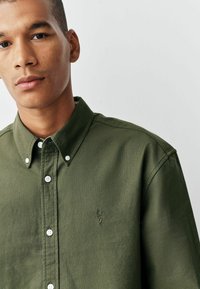 Chemise verte à manches longues en tissu texturé, dotée d'un col pointu, de boutons blancs et d'un petit logo de cerf brodé.