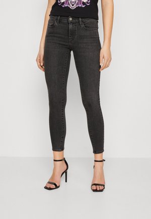Femme portant un jean noir slim et des sandales noires à talons hauts, debout devant un fond clair uni.