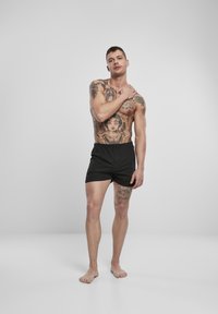 Tetovált férfi modell fekete pamut sortjában, mezítláb áll, egyik kezével a vállán, egy sima szürke háttér előtt.