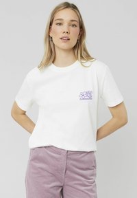 T-shirt en coton blanc avec un motif floral violet sur la poitrine gauche. Manches courtes, col rond et coupe décontractée.