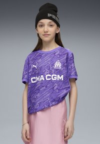 Maglia sportiva viola con motivo testurizzato, loghi bianchi e maniche corte. Indossata con una gonna rosa chiaro e un berretto nero.