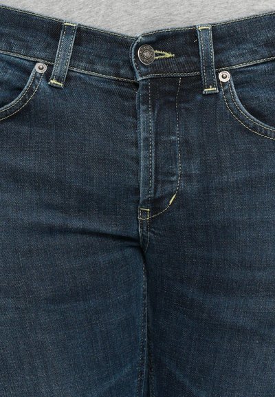 Primo piano di jeans in denim blu scuro con anelli per cintura visibili, rivetti argentati, dettagli di cucitura e una camicia grigia infilata nella vita.