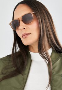 Ochelari de soare rotunzi retro cu rame de culoare bej deschis și lentile colorate, asortați cu o jachetă bomber verde peste un top alb cu gât înalt.