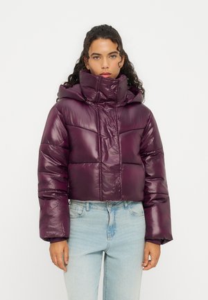 Femme aux cheveux bouclés portant une doudoune brillante violet foncé et un jean bleu clair, debout devant un fond uni.