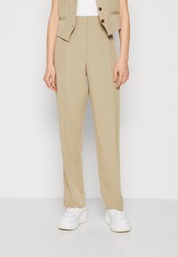 Personne portant un pantalon droit beige, des baskets blanches et un gilet beige sur fond blanc uni.