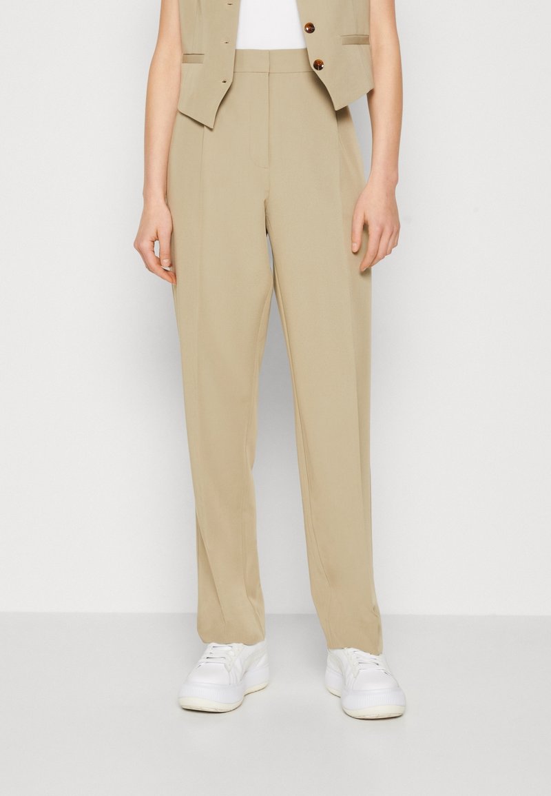 Person som bär beige raka byxor, vita sneakers och en beige väst mot en ren vit bakgrund.