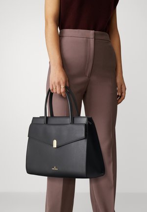 Black leather handbag with a structured design, gold hardware accent, and top handles. Worn by a person in taupe trousers.