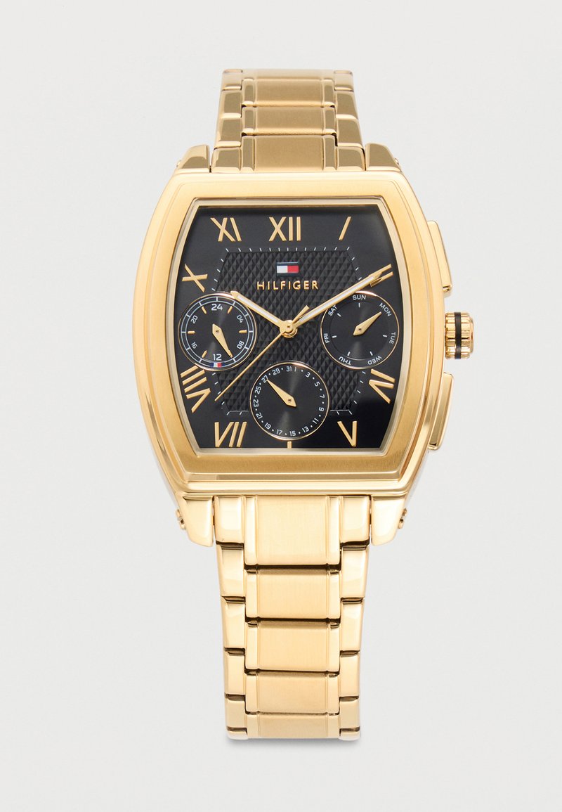 Reloj de pulsera en tono dorado con esfera negra, números romanos, tres subesferas para 24 horas, día y fecha, y el logo de Tommy Hilfiger en el centro superior.