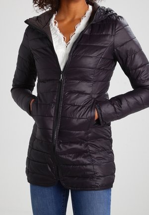 Mujer con chaqueta negra acolchada con capucha sobre un top blanco con ribete de encaje y vaqueros azules, con las manos en los bolsillos de la chaqueta.