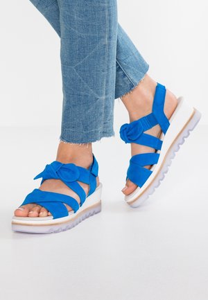 Niebieskie zamszowe sandały z wieloma paskami i detalem w postaci kokardy, podwyższona biała podeszwa na koturnie z teksturowaną szaro-skalną podeszwą. Modny i żywy design.