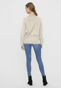 Pull léger beige avec des manches longues et un large décolleté, associé à un jean skinny bleu et des bottines noires. Texture et design simples.