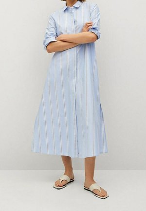 Femme en longue robe bleu clair boutonnée avec des rayures verticales blanches, manches retroussées et sandales beige à boucle pour les orteils, debout les bras croisés.