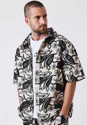 Homme aux cheveux courts et à la barbe, portant une chemise à manches courtes avec des motifs floraux noirs sur un t-shirt blanc, regardant sur le côté.