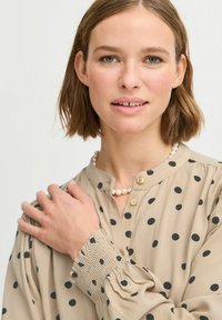 Beige blůza s černými puntíky, s vysokým límečkem, zapínáním na knoflíky a nařasenými manžetami. Perlový náhrdelník jako doplněk.