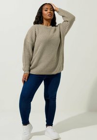 Beige stickad tröja med lös passform och ribbad textur, i kombination med smala blå jeans och vita sneakers.