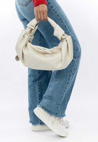 Kremowa, miękka torba typu slouchy z węzłowymi rączkami i zapięciem na zamek. Noszona z niebieskimi dżinsami i białymi trampkami.