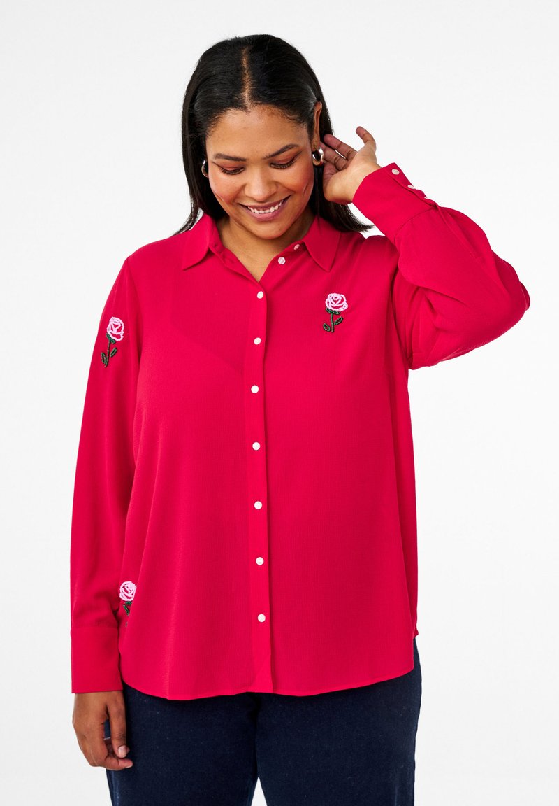 Fuksijas krāsas krekls ar pogām, izgatavots no gludas auduma, ar izšūtām rozā rožu detaļām uz katras piedurknes un klasisku apkakles dizainu.