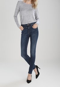 Mujer vestida con una camiseta de manga larga gris, jeans ajustados oscuros y tacones altos negros, de pie con las manos en los bolsillos y las piernas cruzadas.