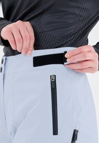 Ljusblå skidbyxor med ett kardborreband i midjan, dragkedjefickor och en slät yta. Har svarta detaljer och en diskret varumärkesaccent.