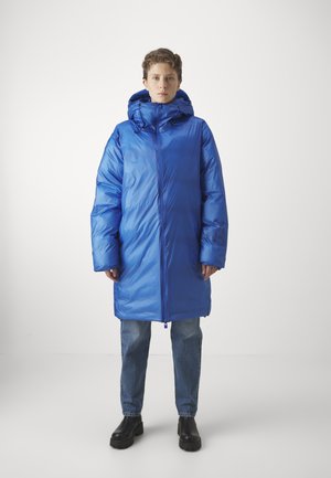 Puffet blå parka med hætte, lavet af skinnende, glat stof. Lange ærmer og lynlås, parret med afslappede blå jeans og sorte støvler.