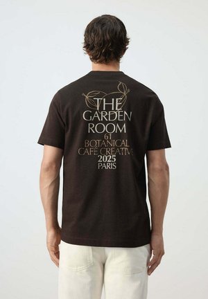 Hombre que lleva una camiseta negra con "The Garden Room 61 Botanical Café Creative 2025 Paris" impreso en la espalda, combinado con pantalones claros.