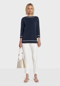 Haut en maille bleu marine avec accents blancs, manches trois-quarts ; associé à un pantalon blanc sur mesure et un sac à main beige clair. Sandales argentées à lanières.