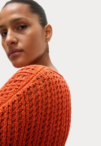 Oranje gehaakte top met een open maasontwerp, Featuring een brede halslijn en een losse pasvorm. De achtergrond is neutraal, waardoor de textuur van het kledingstuk benadrukt wordt.
