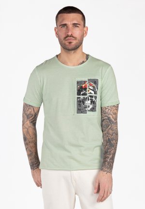 Uomo con tatuaggi che indossa una maglietta verde chiaro con una grafica di un teschio con fiori rossi divisa in quattro quadrati sul petto.