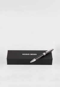Black ballpoint pen with chrome accents, resting on a black box. Box features "HUGO BOSS" in white lettering. Simple, sleek design.