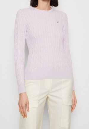 Femme portant un pull en maille torsadée violet clair et un pantalon écru avec de grandes poches à l'avant, debout devant un fond uni.