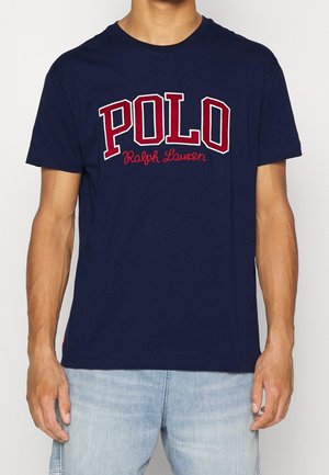 Mężczyzna noszący granatowy t-shirt z dużym napisem „POLO Ralph Lauren” na piersi w kolorze czerwonym i białym oraz jasnoniebieskie dżinsy.