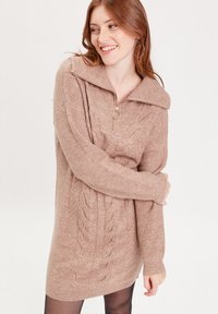 Robe pull en tricot brun clair avec un col zippé, présentant des détails en relief et des poignets côtelés, offrant un look chaleureux et texturé.